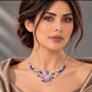 A close-up of a woman wearing a sparkling floral necklace with pink, blue, and purple stones on a silver-toned chain, paired with a satin pink top against a soft gray background.