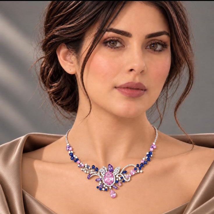 A close-up of a woman wearing a sparkling floral necklace with pink, blue, and purple stones on a silver-toned chain, paired with a satin pink top against a soft gray background.